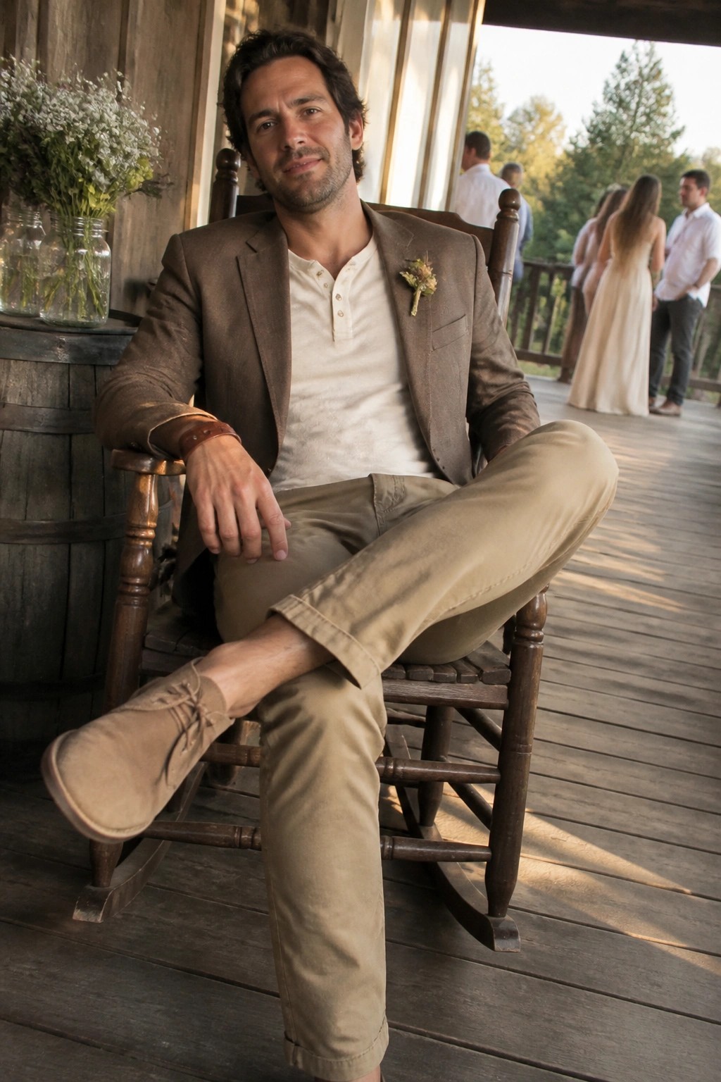 Man seated in wooden rocking chair wearing brown blazer over cream henley shirt with khaki trousers and brown suede shoes plus small boutonniere on lapel.