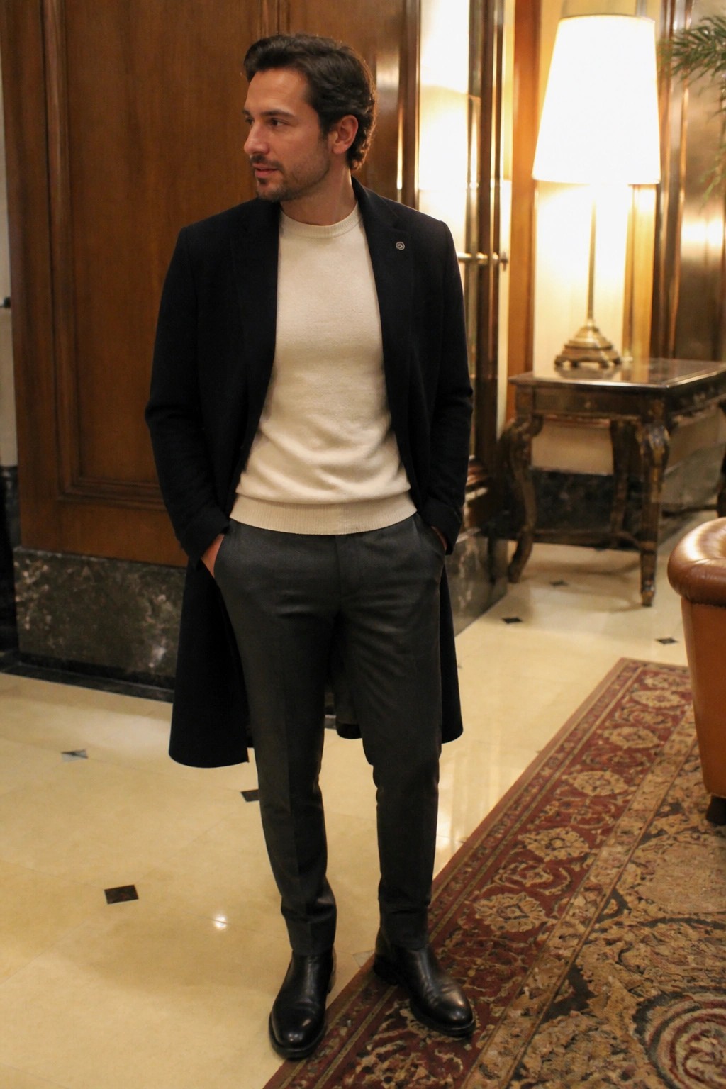 Man wearing black overcoat over cream knit sweater with charcoal formal pants and black dress boots standing indoors with hands in pockets.