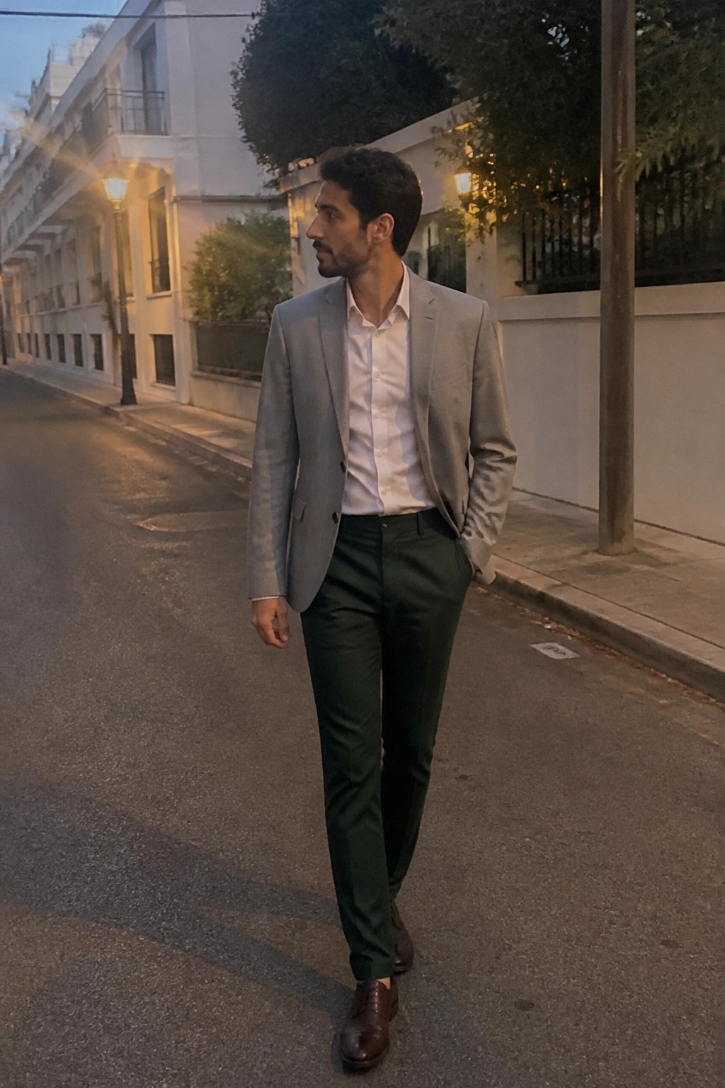 Man wearing a light gray blazer, white dress shirt, dark green formal trousers, and brown leather dress shoes while standing on a city street at dusk.