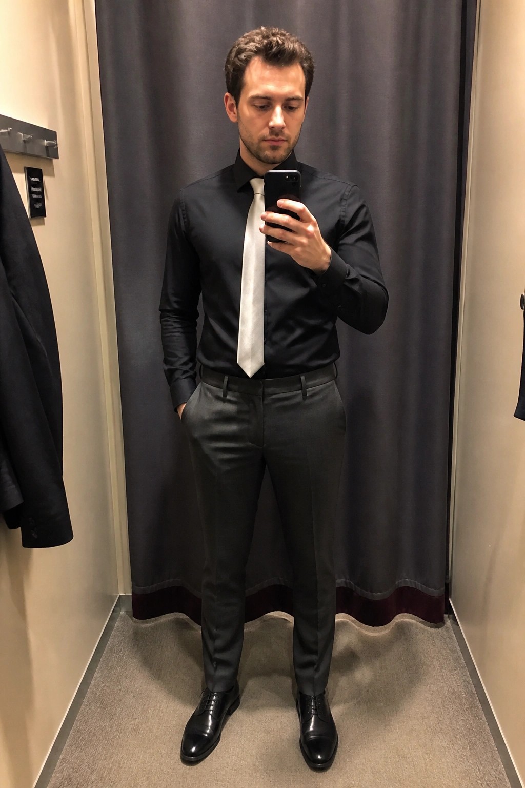Man in black shirt and white tie taking mirror selfie in fitting room