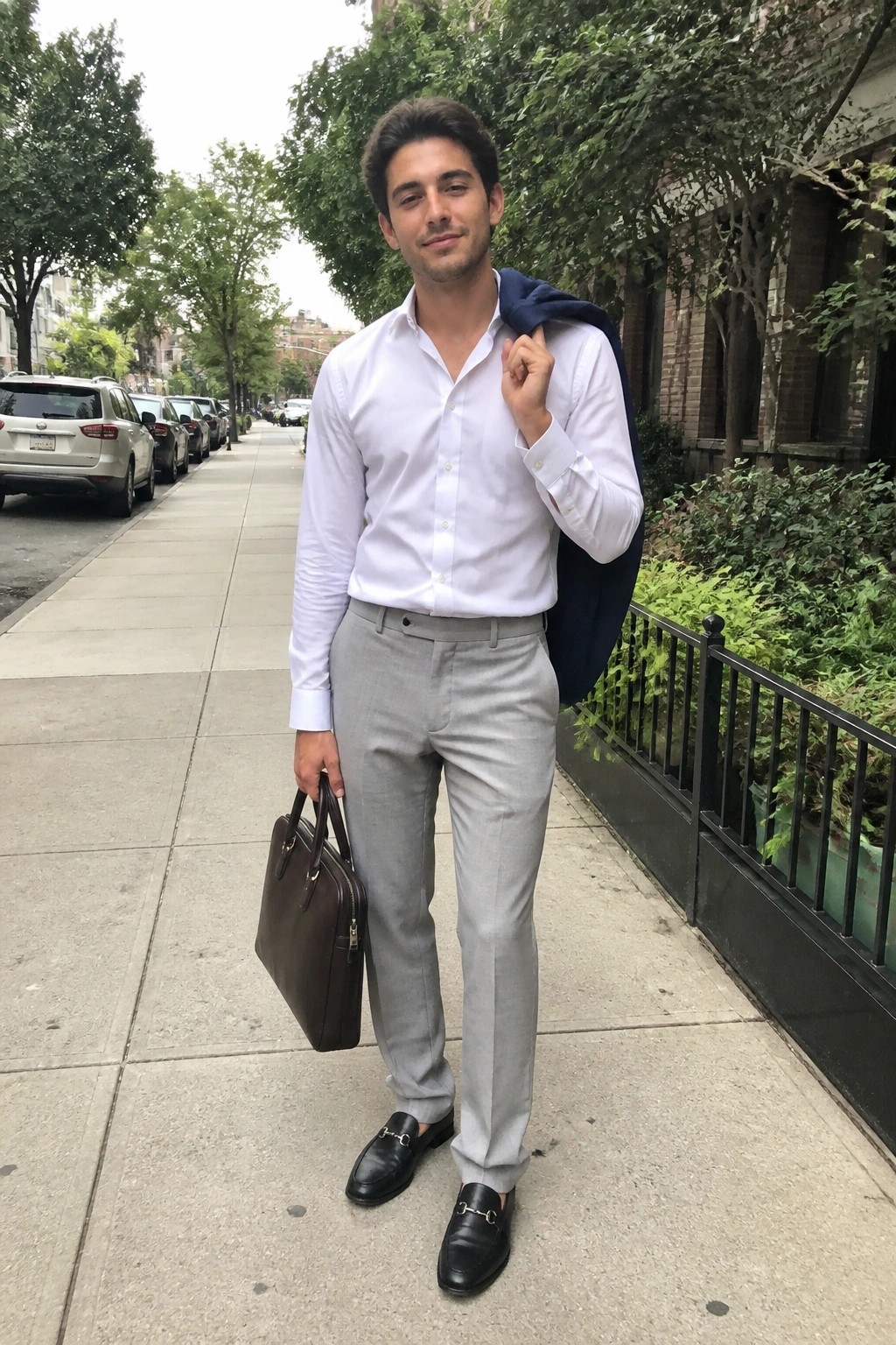 A man in a white button-down shirt tucked into light gray formal pants, paired with black horsebit loafers, holding a brown leather briefcase while a navy blazer rests over one shoulder.