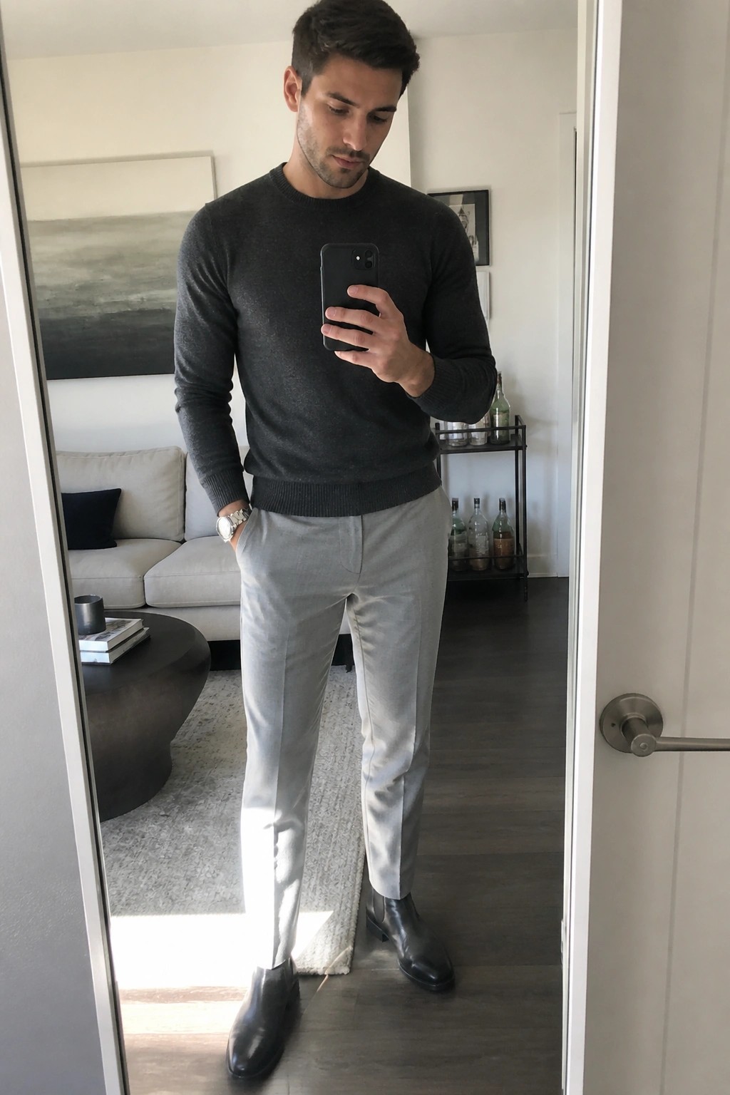 Man in charcoal gray sweater, light gray formal trousers, and black Chelsea boots taking a mirror selfie indoors.