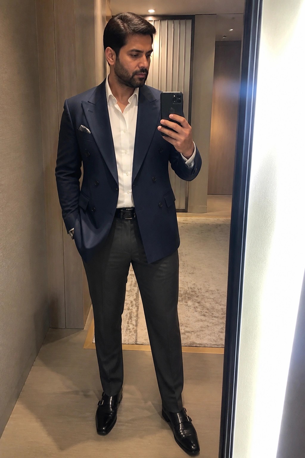 Man wearing a navy double breasted blazer over a white dress shirt with grey formal pants and black double monk strap shoes while taking a mirror selfie.
