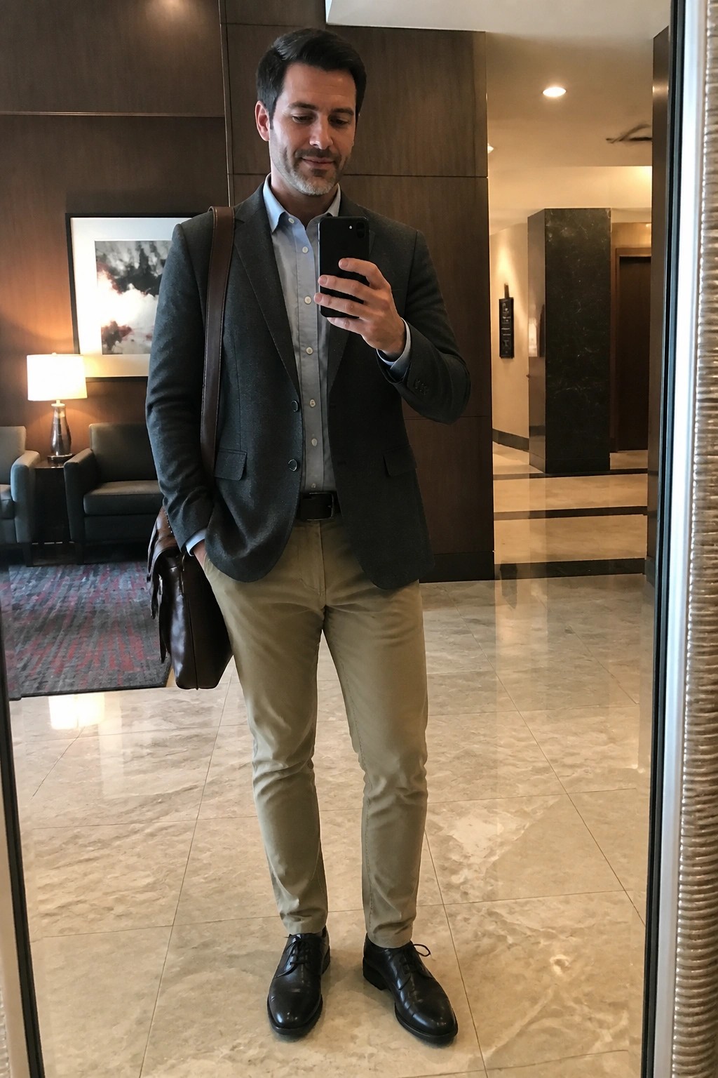 Man in gray blazer and khaki pants taking mirror selfie in hotel lobby
