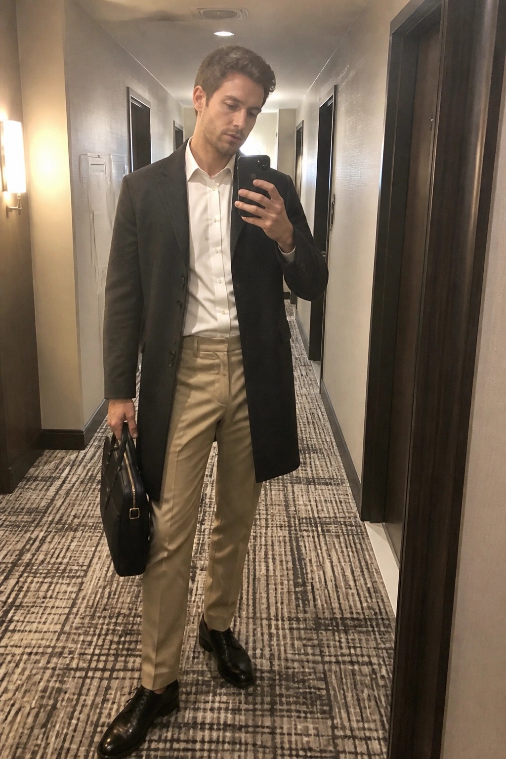 Man in dark overcoat and beige pants taking mirror selfie in hallway