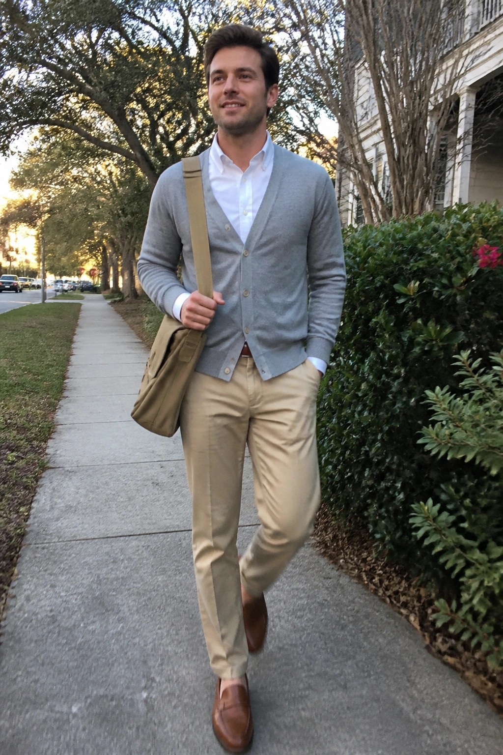 Man in gray cardigan and khaki pants walks sidewalk with tan messenger bag