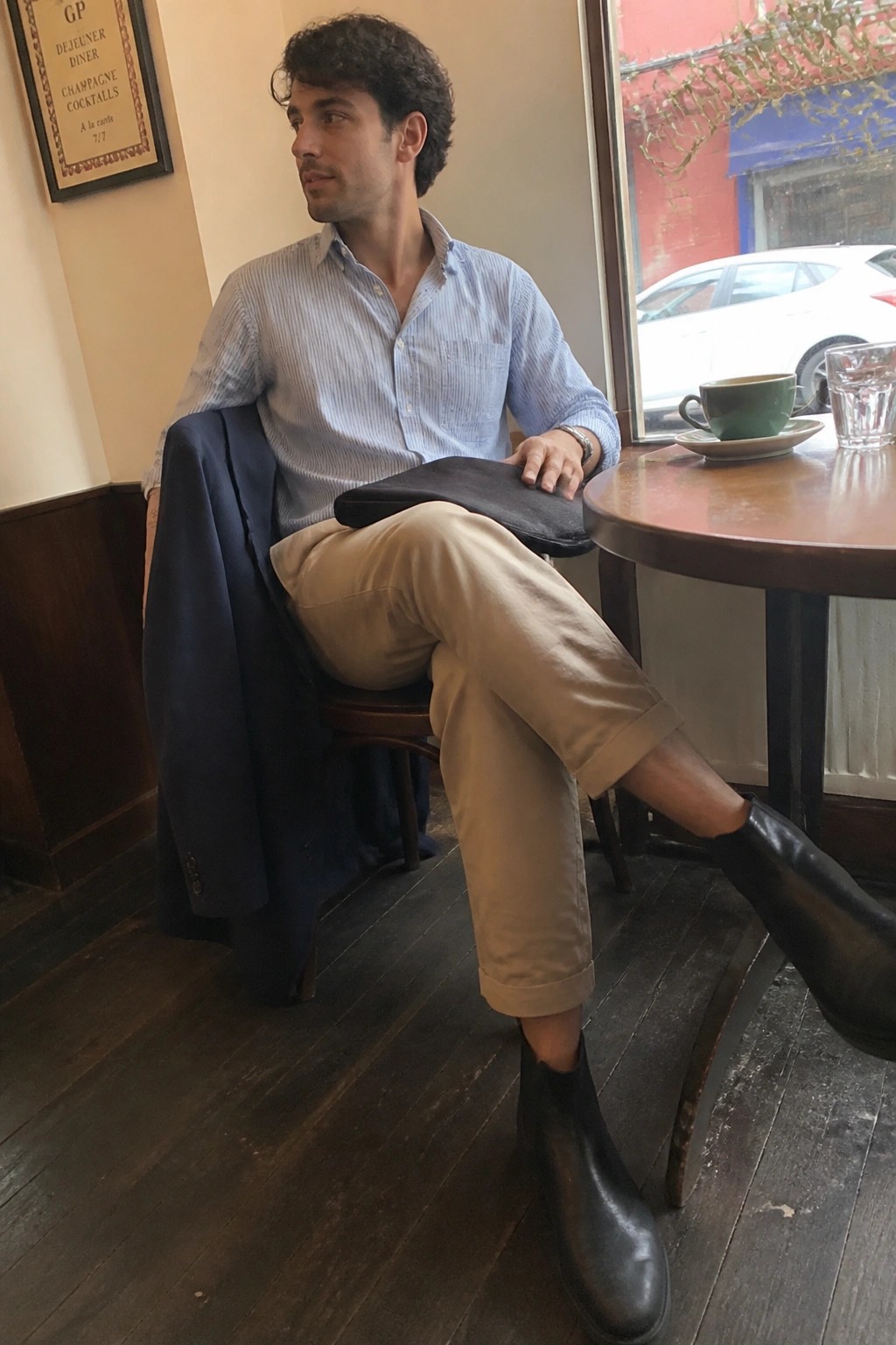 Man seated in a cafe wearing light blue striped button down shirt, khaki pants, navy jacket draped nearby, black Chelsea boots, and holding a black bag on his lap.