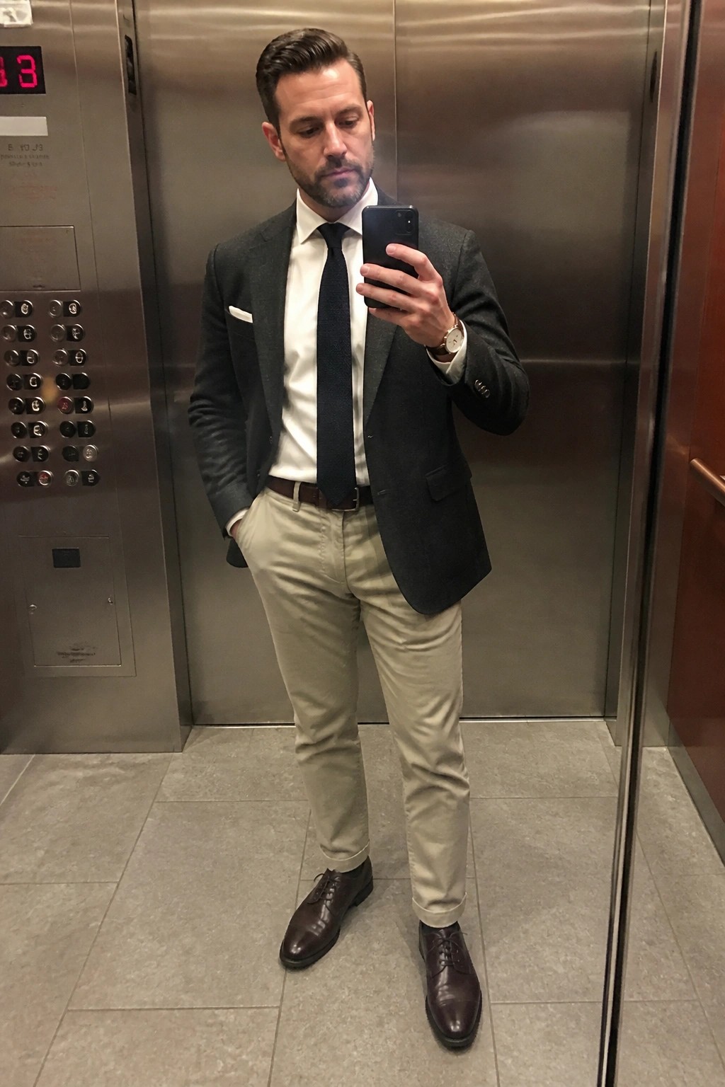 Man in gray blazer, white shirt, tie, and khaki pants taking elevator selfie.