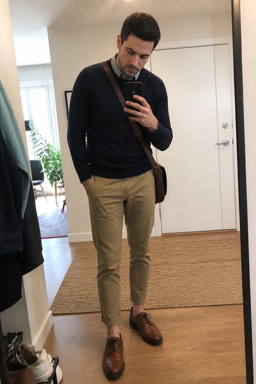 Man in navy sweater and khaki pants taking mirror selfie with brown bag