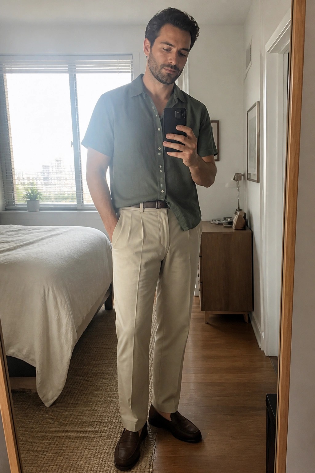 Man in sage green shirt and beige trousers taking mirror selfie in bedroom