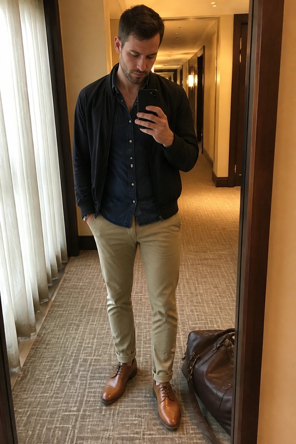 Man in navy jacket and khaki pants taking mirror selfie beside leather bag in hallway