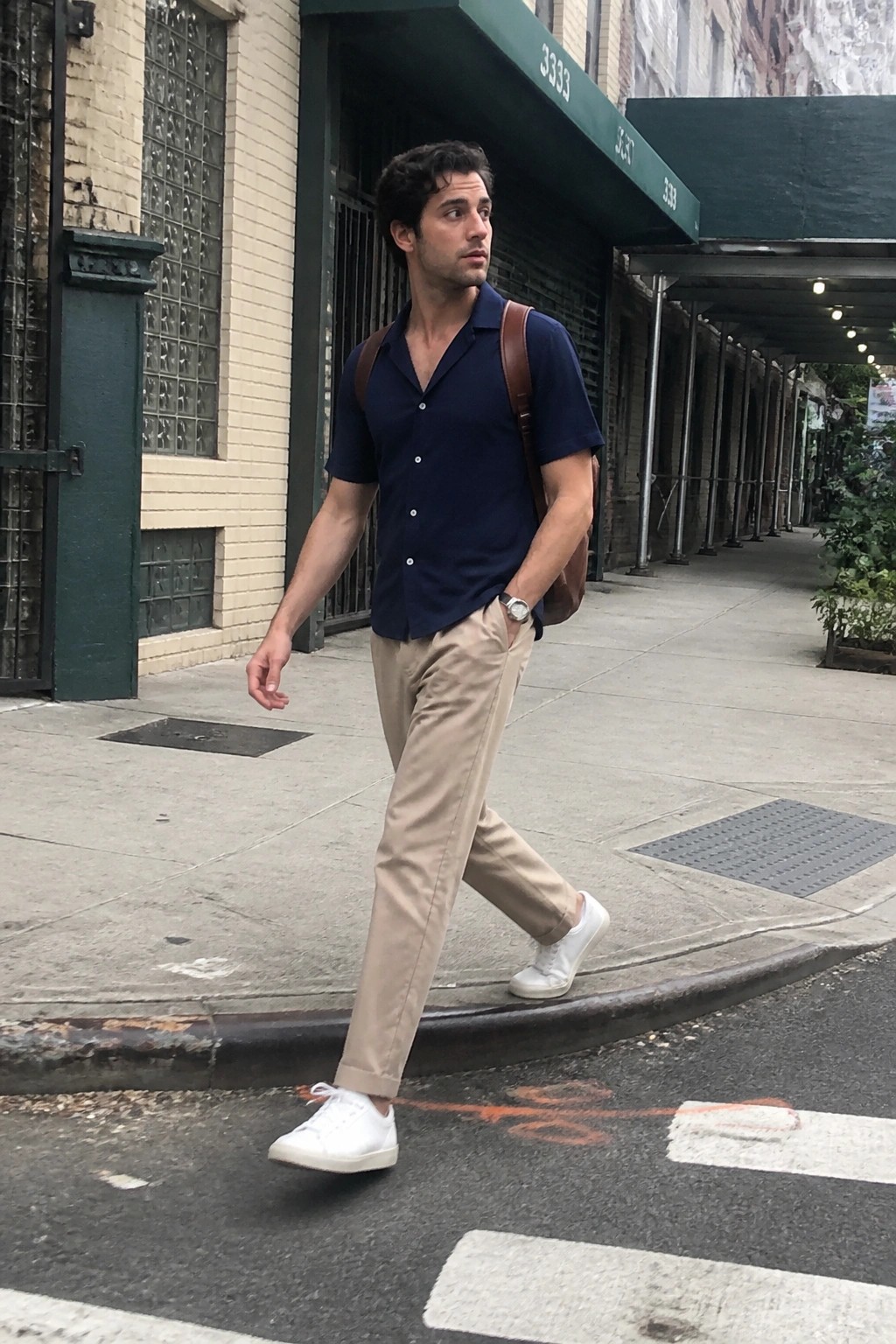 Man in navy shirt and beige pants crossing city street with brown leather bag