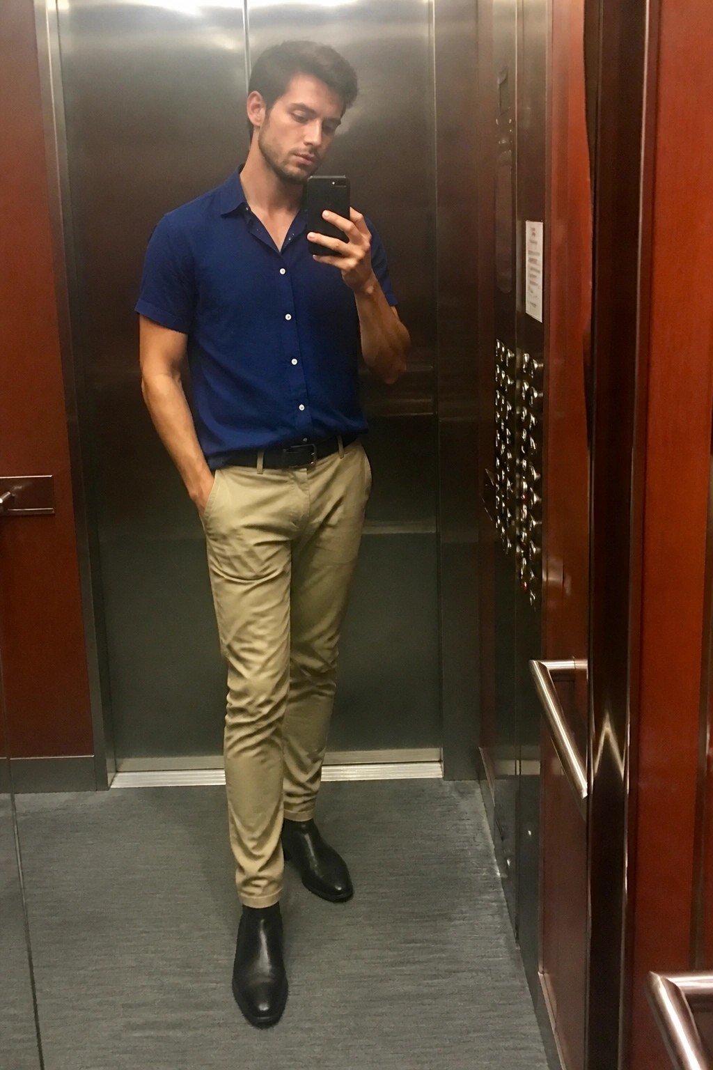 Man in blue shirt and khaki pants taking mirror selfie in elevator