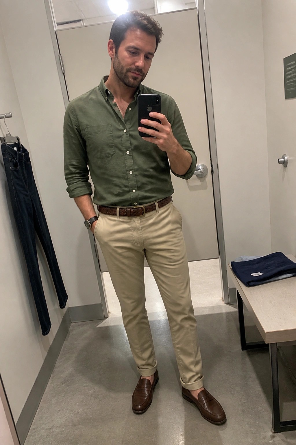Man in green shirt and khaki pants taking mirror selfie in fitting room