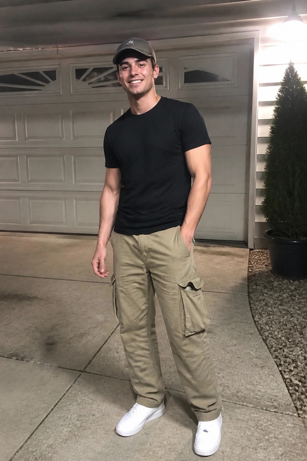 Smiling man in black t-shirt, khaki cargo pants, white sneakers, and cap by garage.