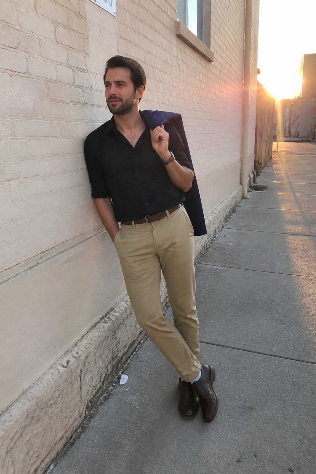 Man in black shirt and khakis holding navy blazer against brick wall at sunset