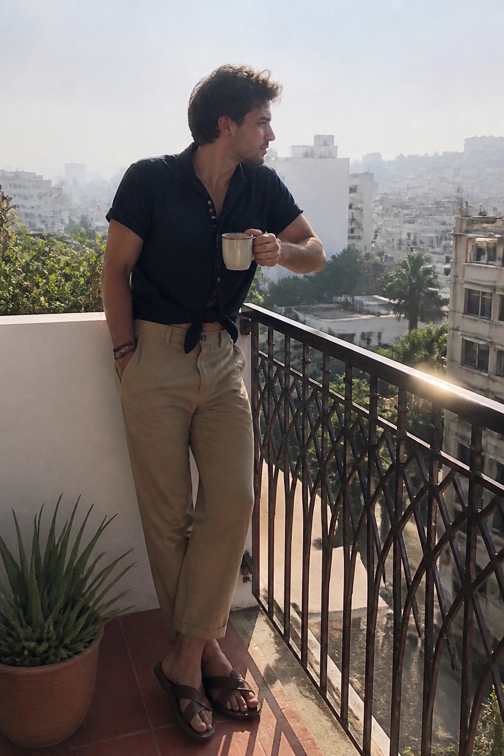 Man in tied navy shirt and khaki pants holding mug on sunny city balcony