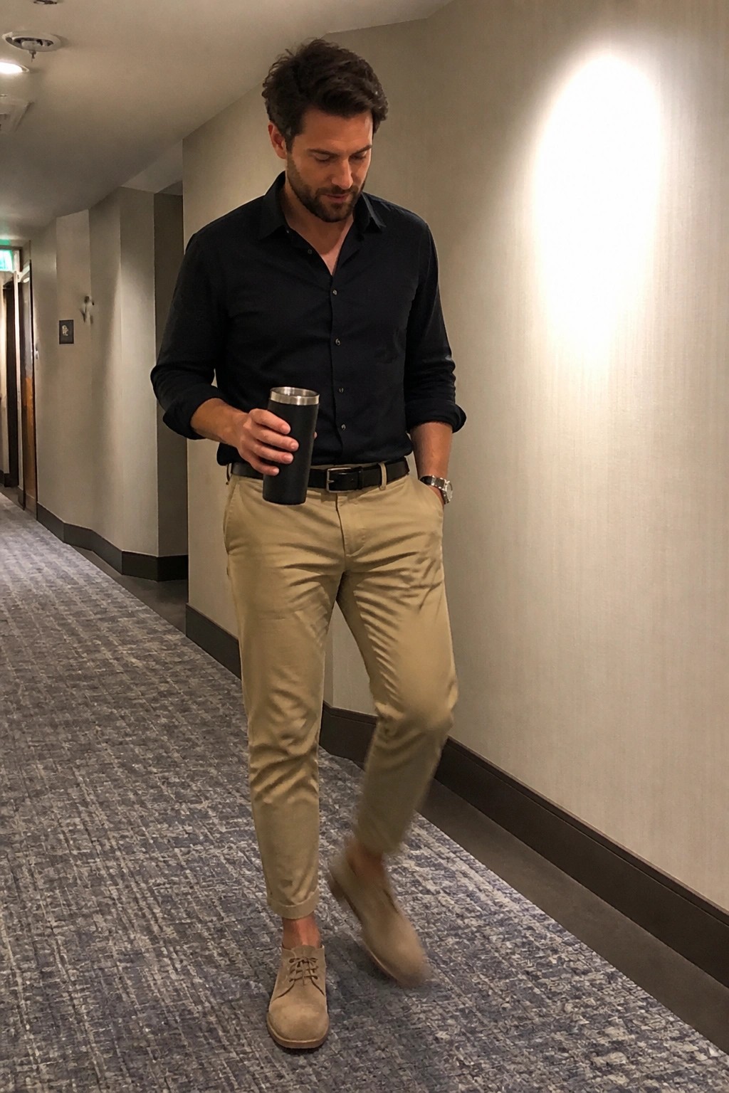 Man in navy shirt and khaki pants walking hallway holding black tumbler