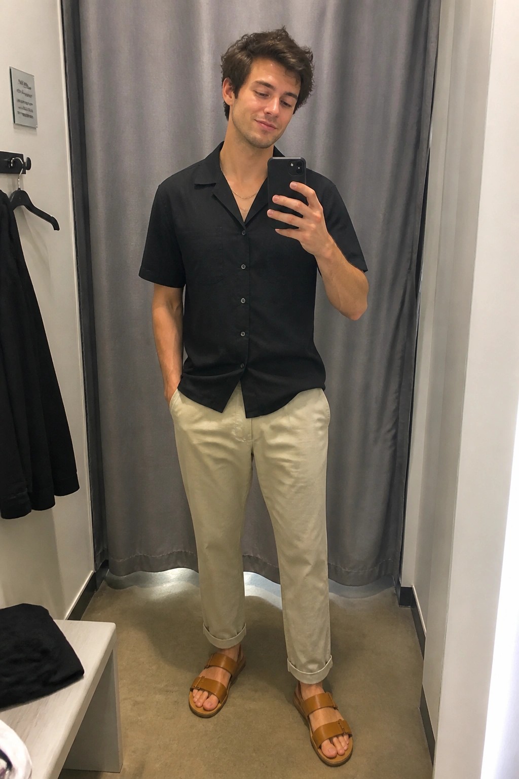 Young man in black shirt and beige pants taking mirror selfie in fitting room