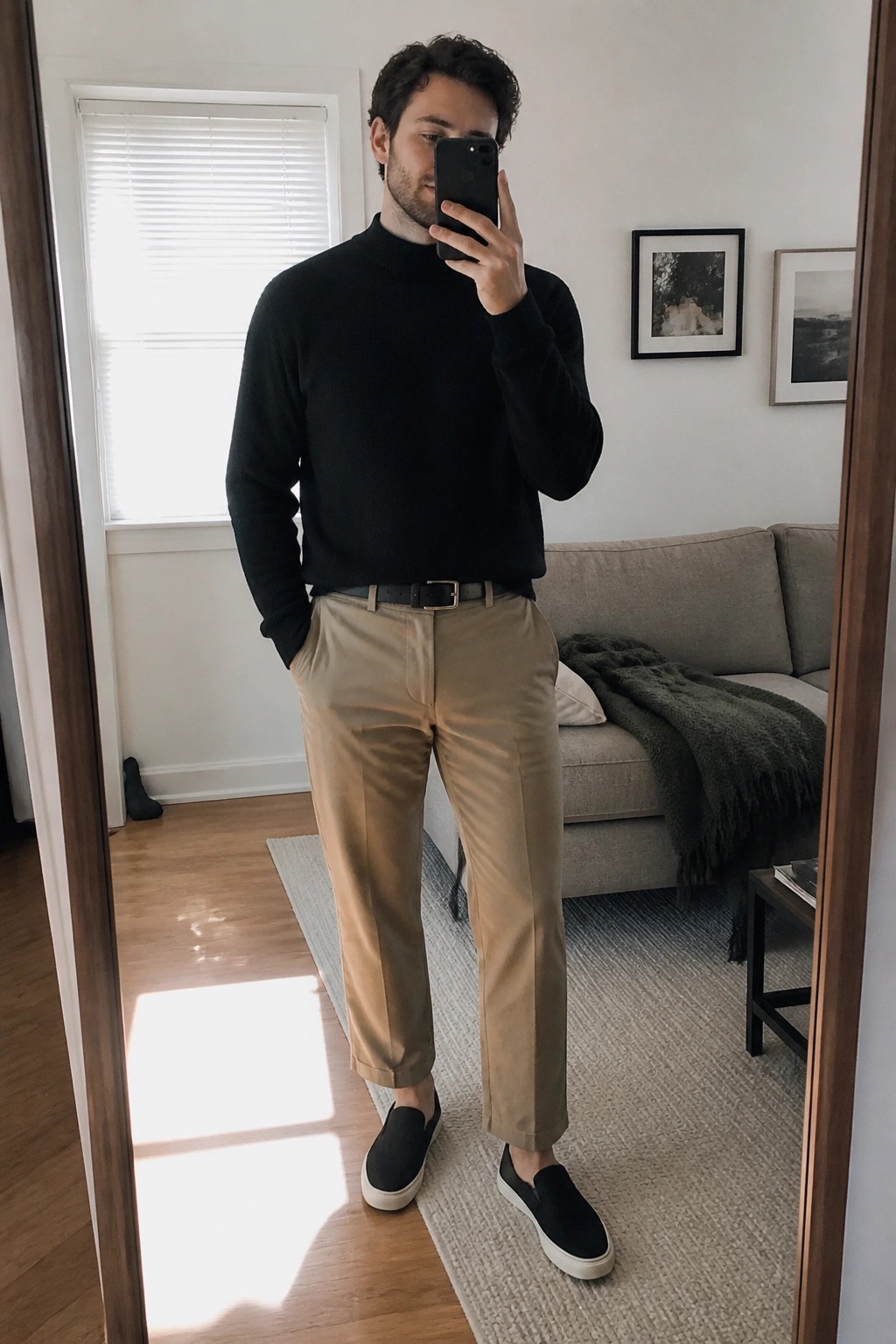 Man in black turtleneck and khaki pants taking mirror selfie in living room.