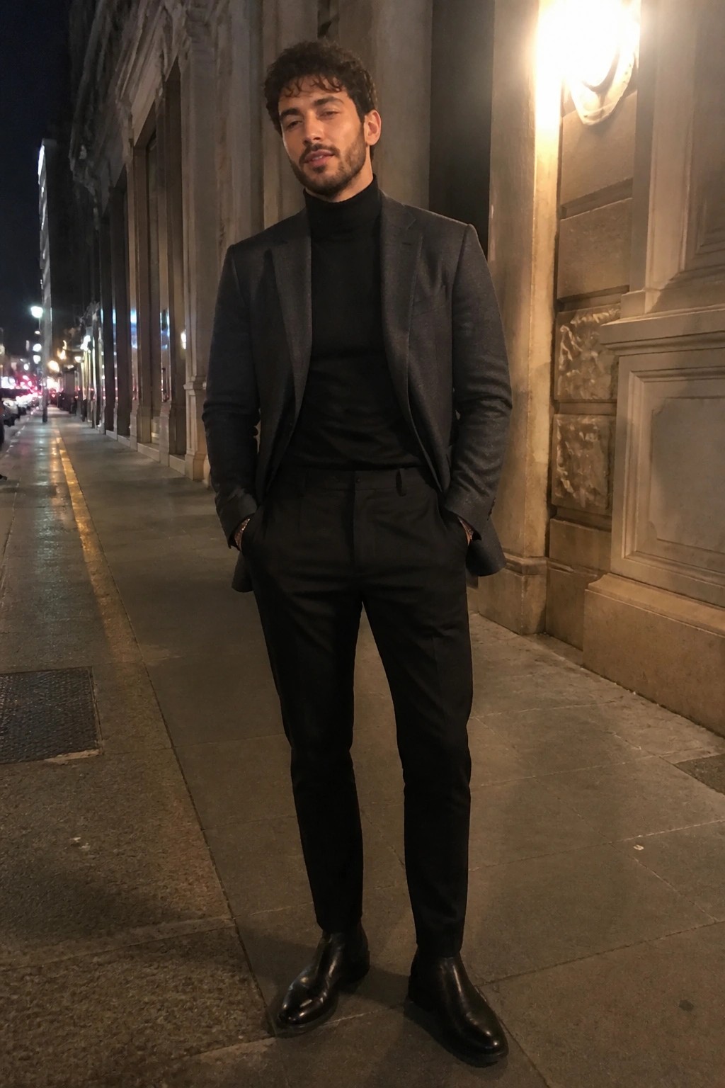 Man in black suit and turtleneck stands on nighttime city sidewalk, hands in pockets.