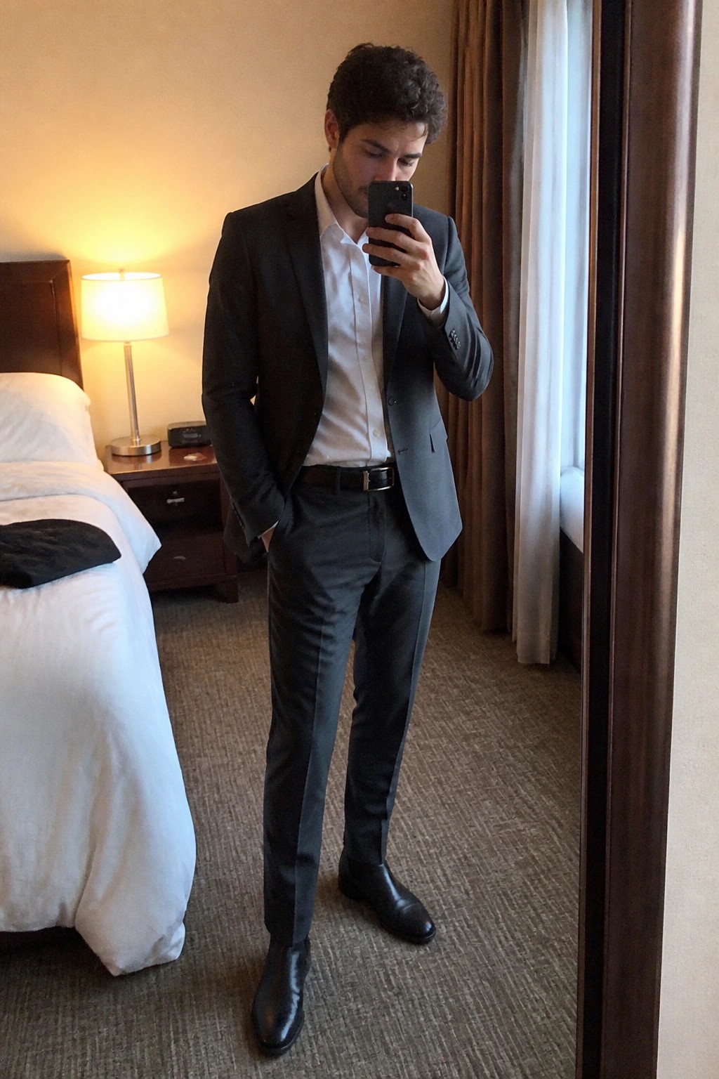 Man in gray suit taking mirror selfie in hotel room with white bed