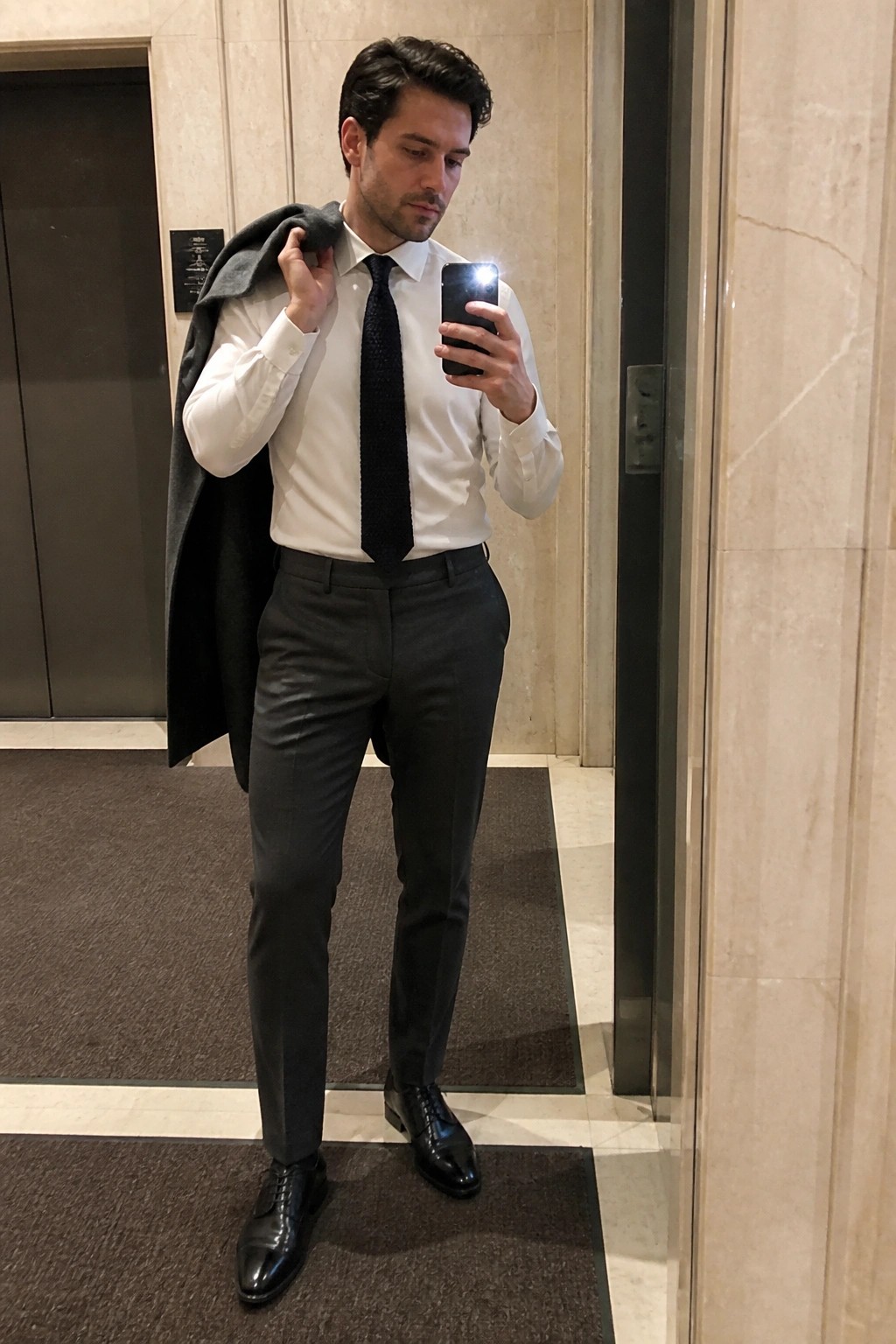 Man in white shirt and black tie holding coat while taking phone selfie