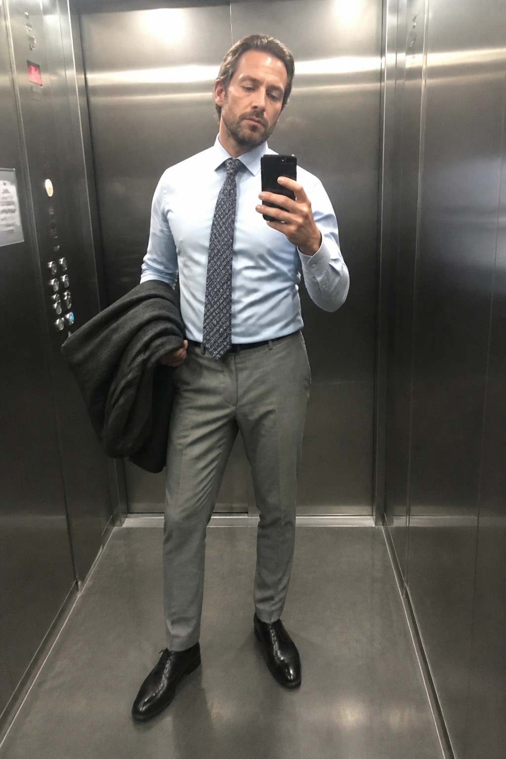 Man in blue shirt and gray pants taking phone selfie in elevator holding jacket