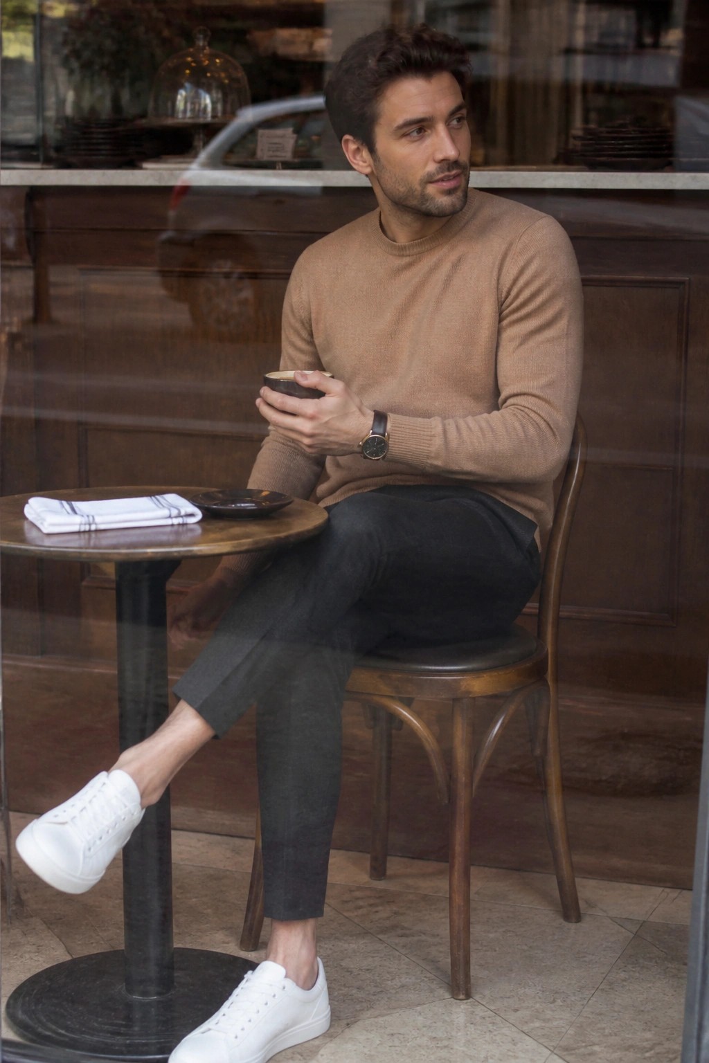 Man in tan sweater holds cup at cafe table wearing white sneakers