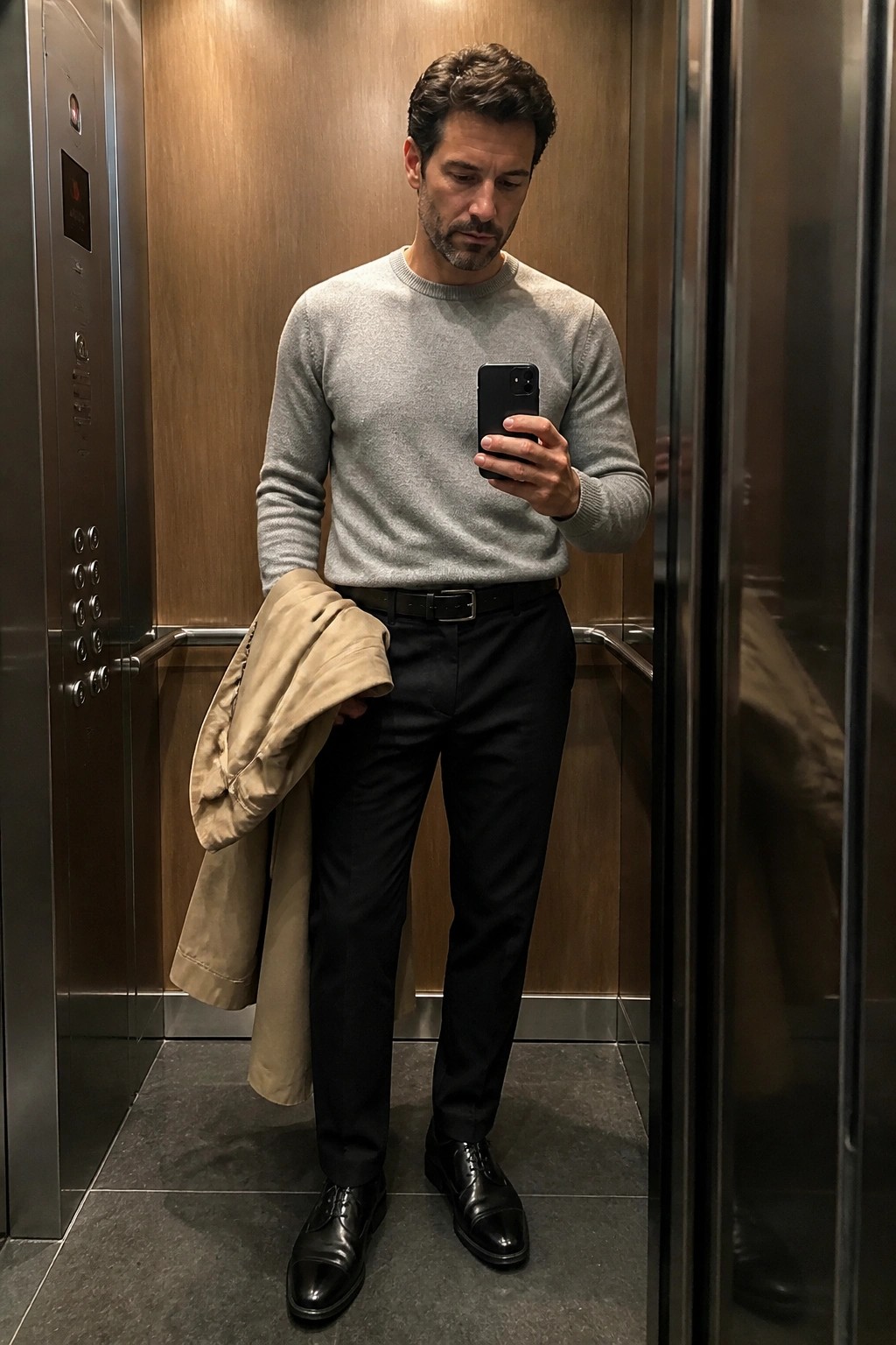 Man in gray sweater holding beige coat takes mirror selfie in elevator.