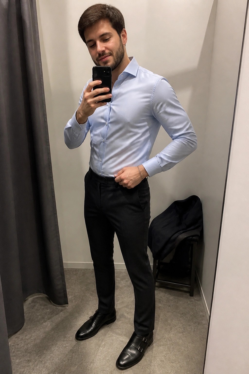 Man wearing light blue button down shirt tucked into black formal trousers with black double monk strap shoes taking a mirror selfie in a fitting room.