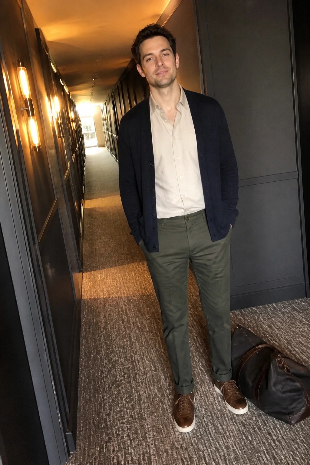 Man in beige shirt, navy cardigan, and green pants in hallway with duffel bag