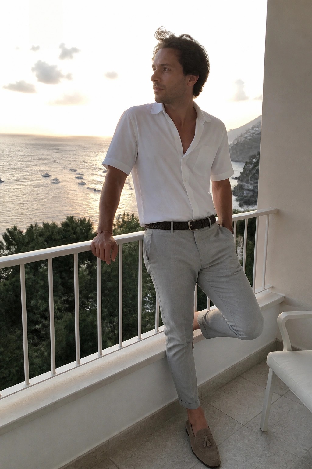 Man in white shirt and gray pants posing on balcony overlooking sea at sunset