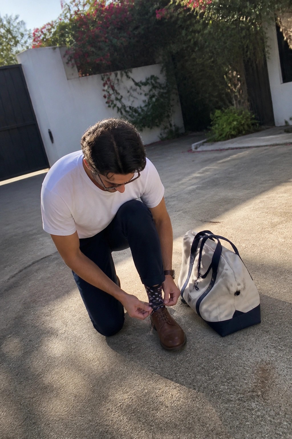 Man in white t-shirt tying brown shoe beside beige duffel bag on pavement