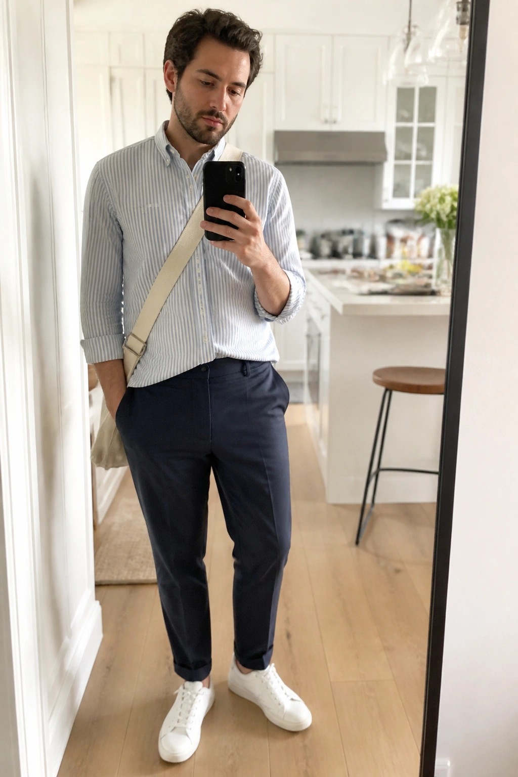 Man in striped shirt and navy pants taking mirror selfie in kitchen.