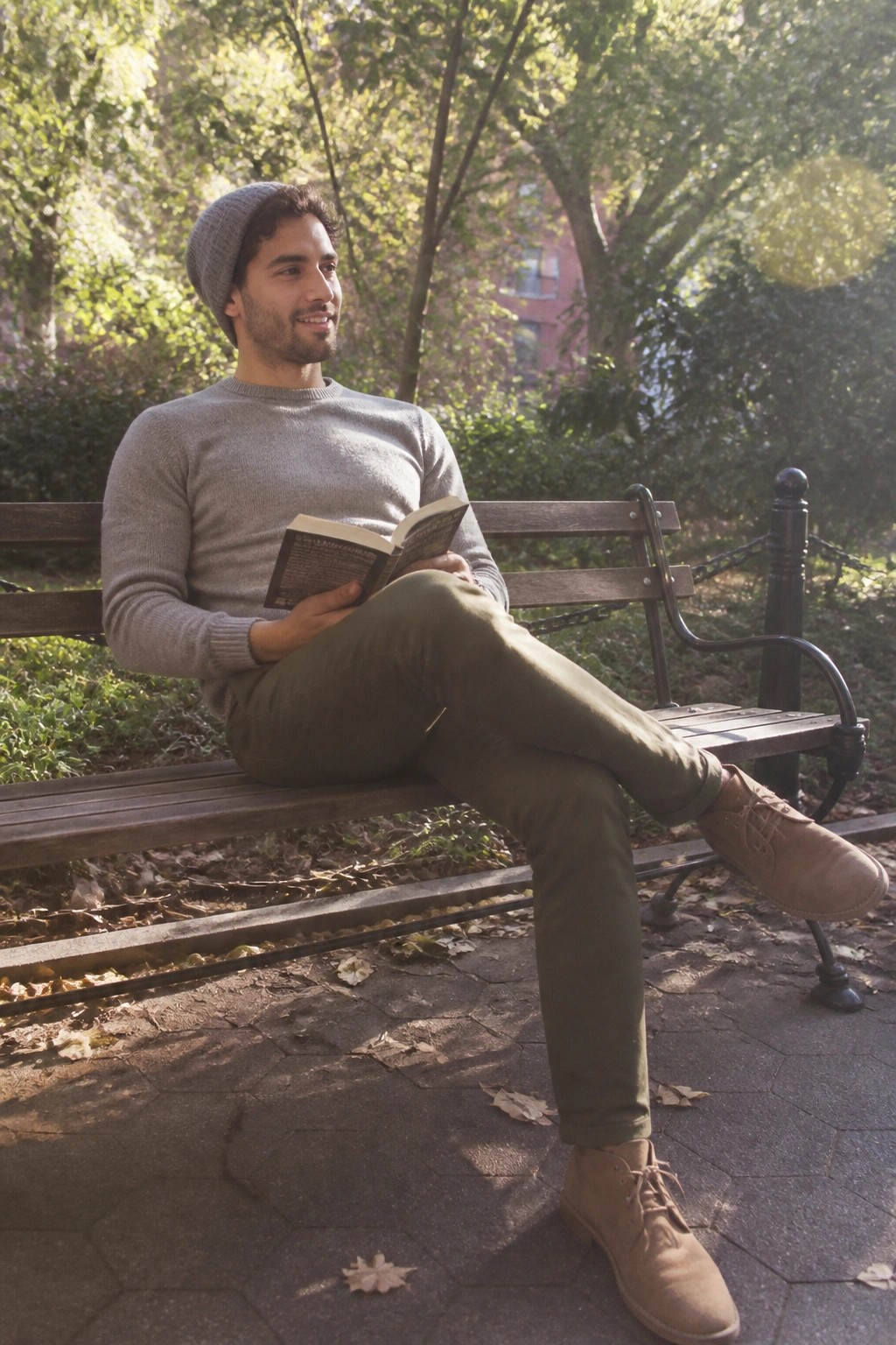 Man in gray sweater and beanie reading open book on wooden park bench.