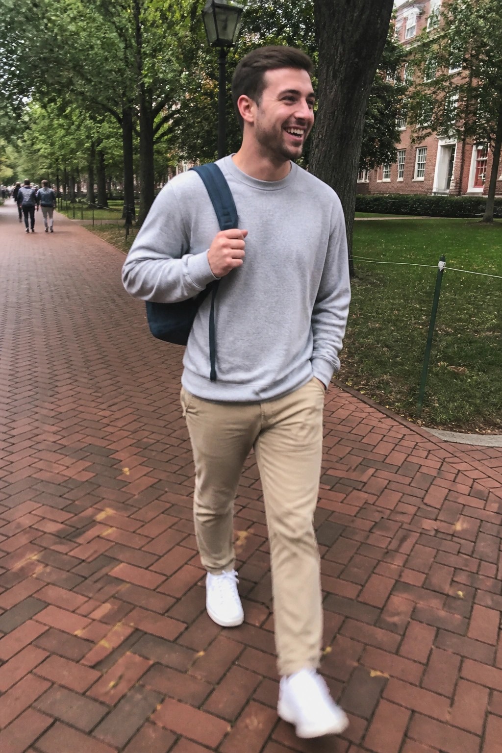 A man wearing a gray crewneck sweatshirt, beige khaki pants, white sneakers, and a blue backpack slung over one shoulder while standing on a brick walkway.