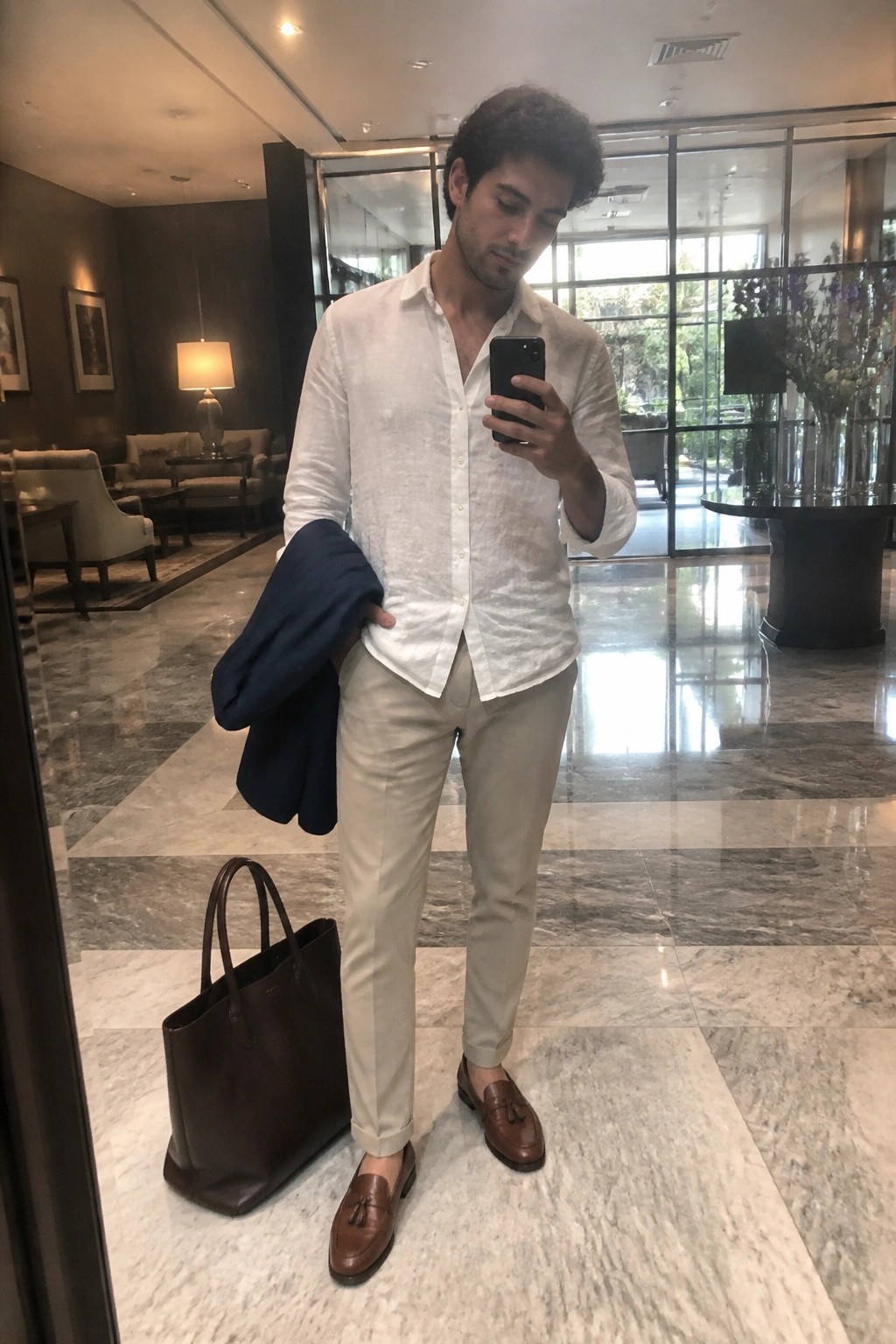 Man in white shirt and beige pants holds phone with navy jacket near brown tote.