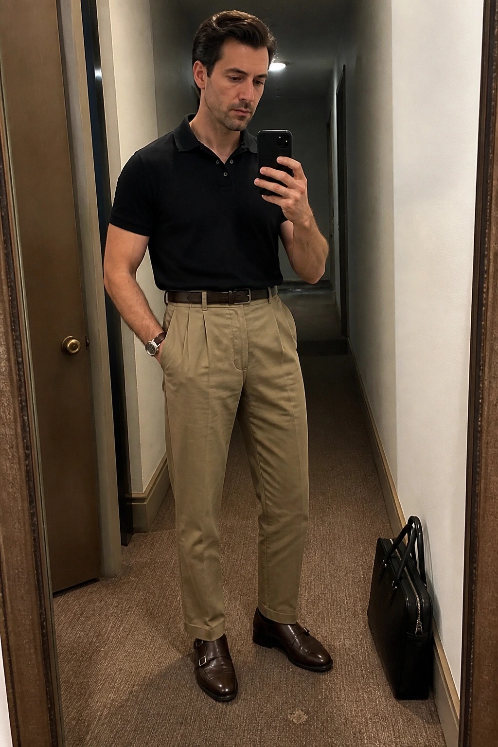 Man in black polo and khaki pants taking mirror selfie beside black briefcase.