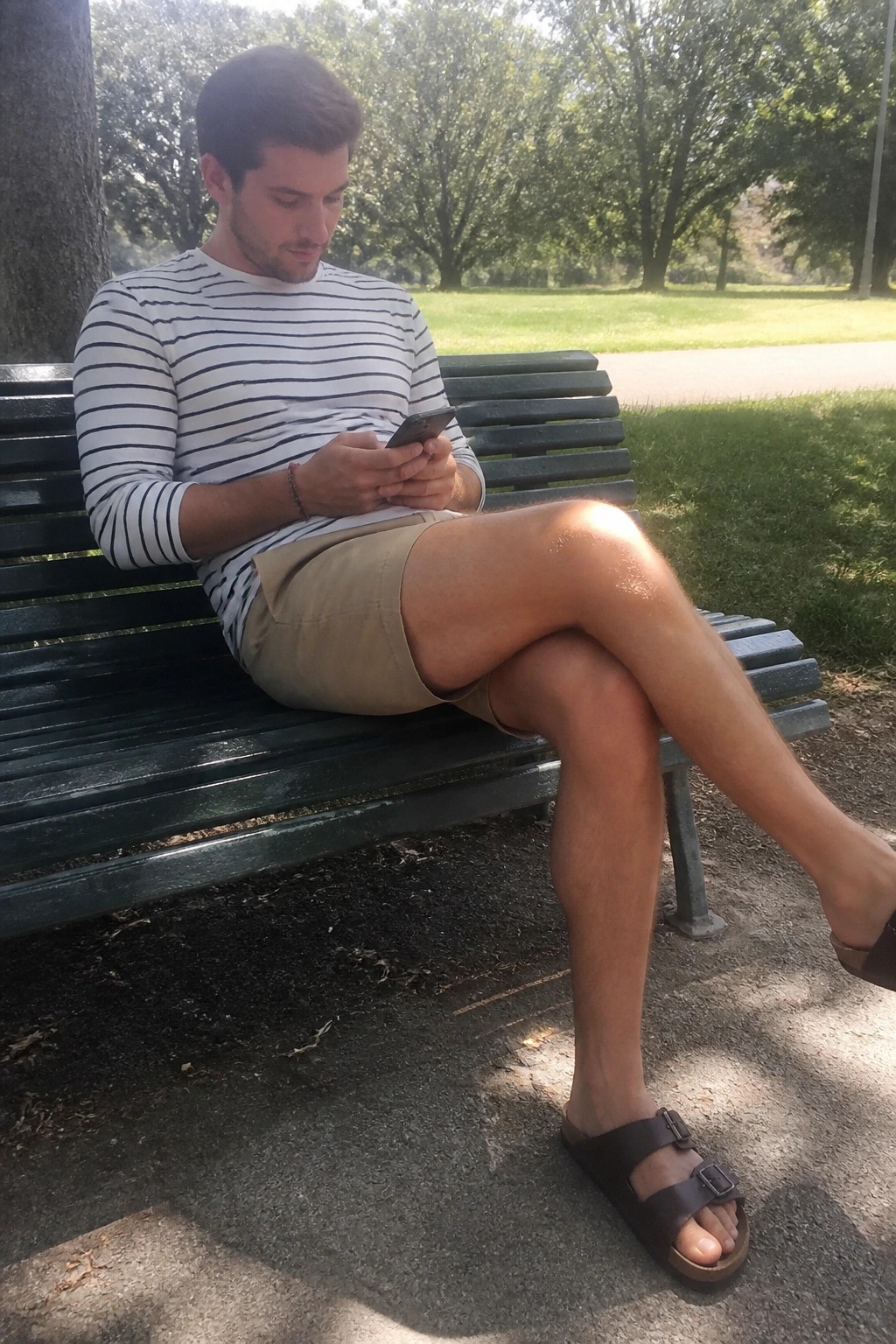 Man in striped shirt and shorts checks phone on green park bench