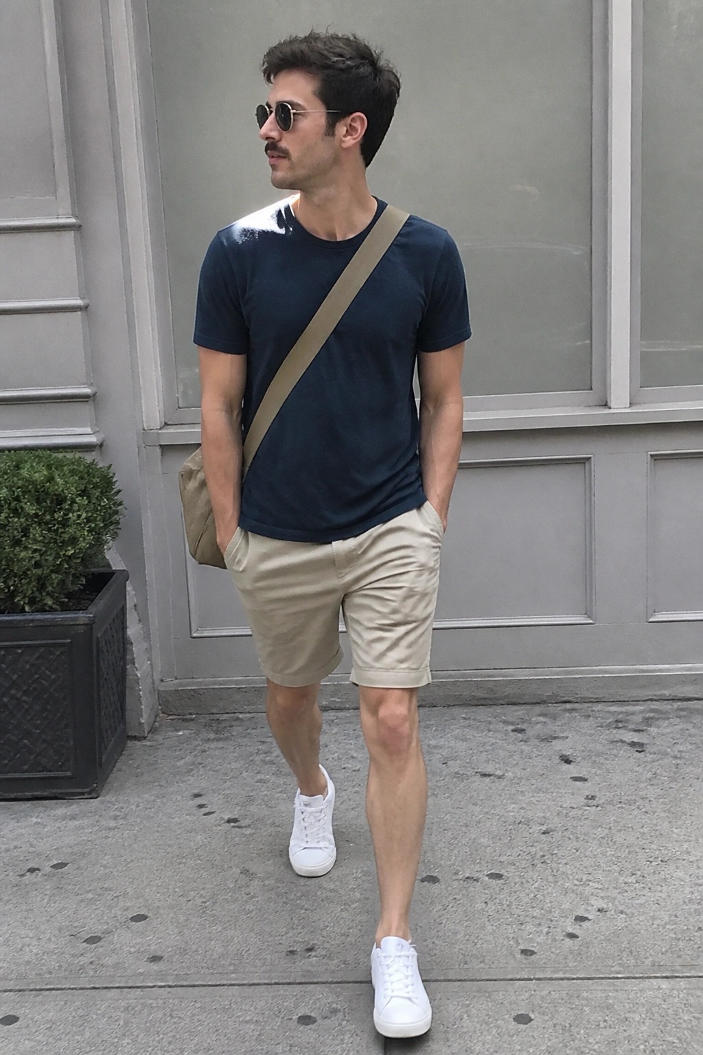Man in navy t-shirt and khaki shorts with beige crossbody bag on sidewalk