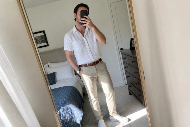 Man in white short-sleeve shirt and khaki pants taking bedroom mirror selfie.