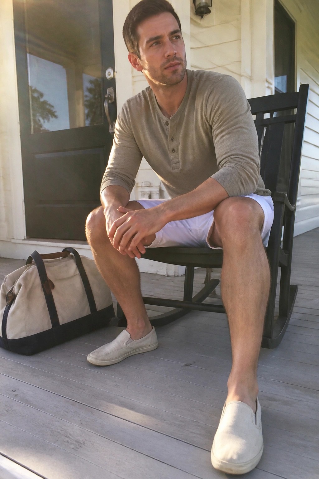 Man seated on a porch wearing a khaki henley shirt, white shorts, and light canvas slip-on shoes with a canvas bag nearby.
