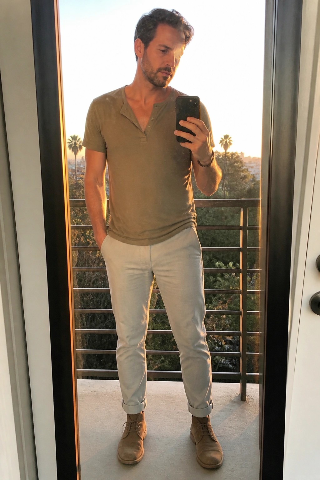 Man in olive henley and beige pants taking balcony mirror selfie at sunset