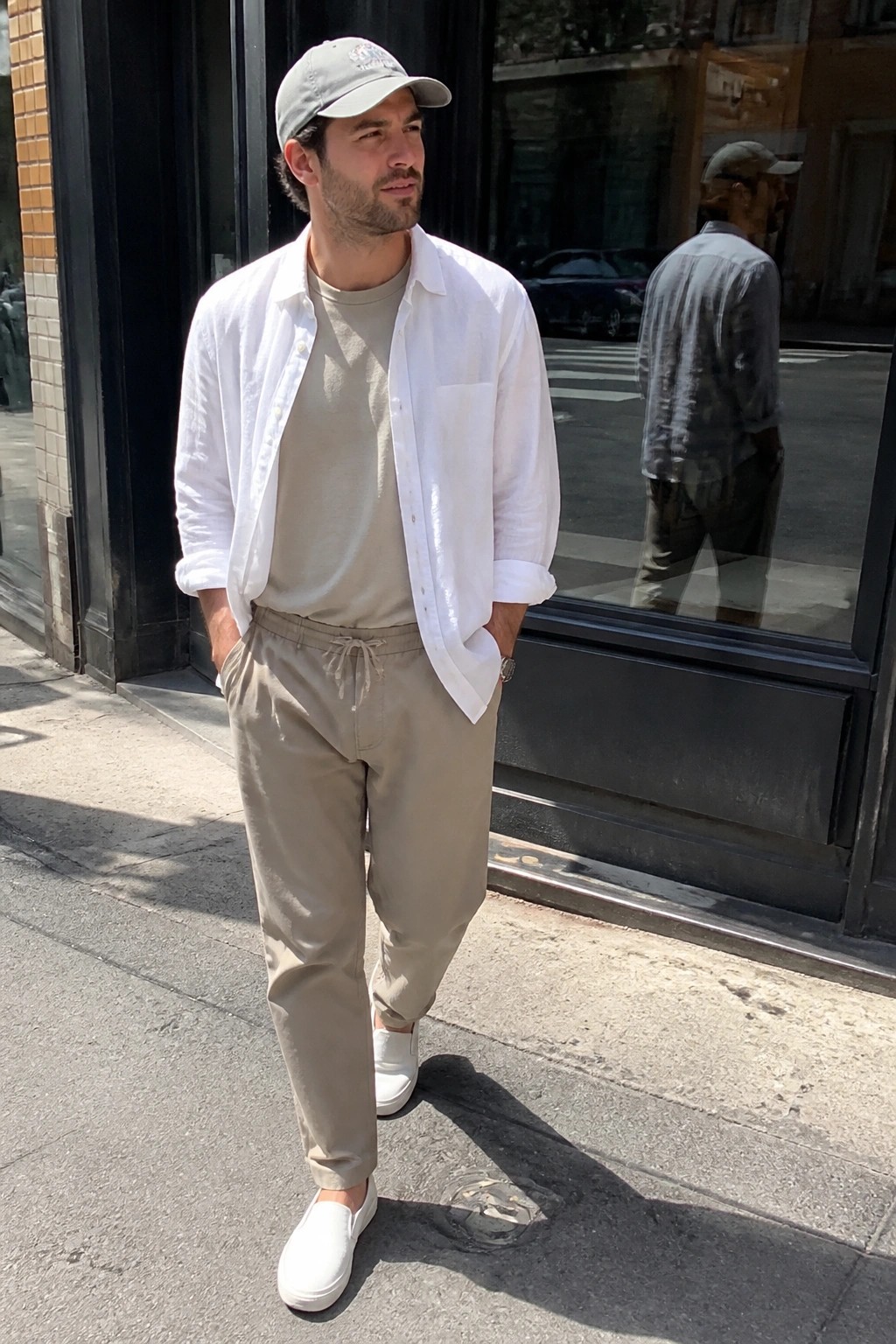 Man wearing open white long sleeve shirt over beige crewneck tee with khaki drawstring pants and white slip on sneakers plus light gray cap standing on sidewalk.