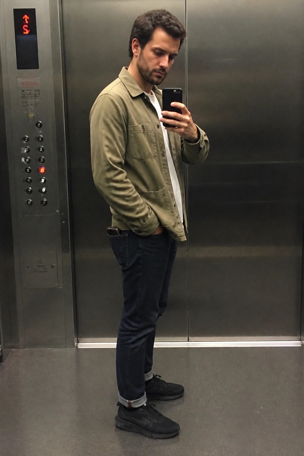 Man in green shirt and jeans taking mirror selfie in elevator.