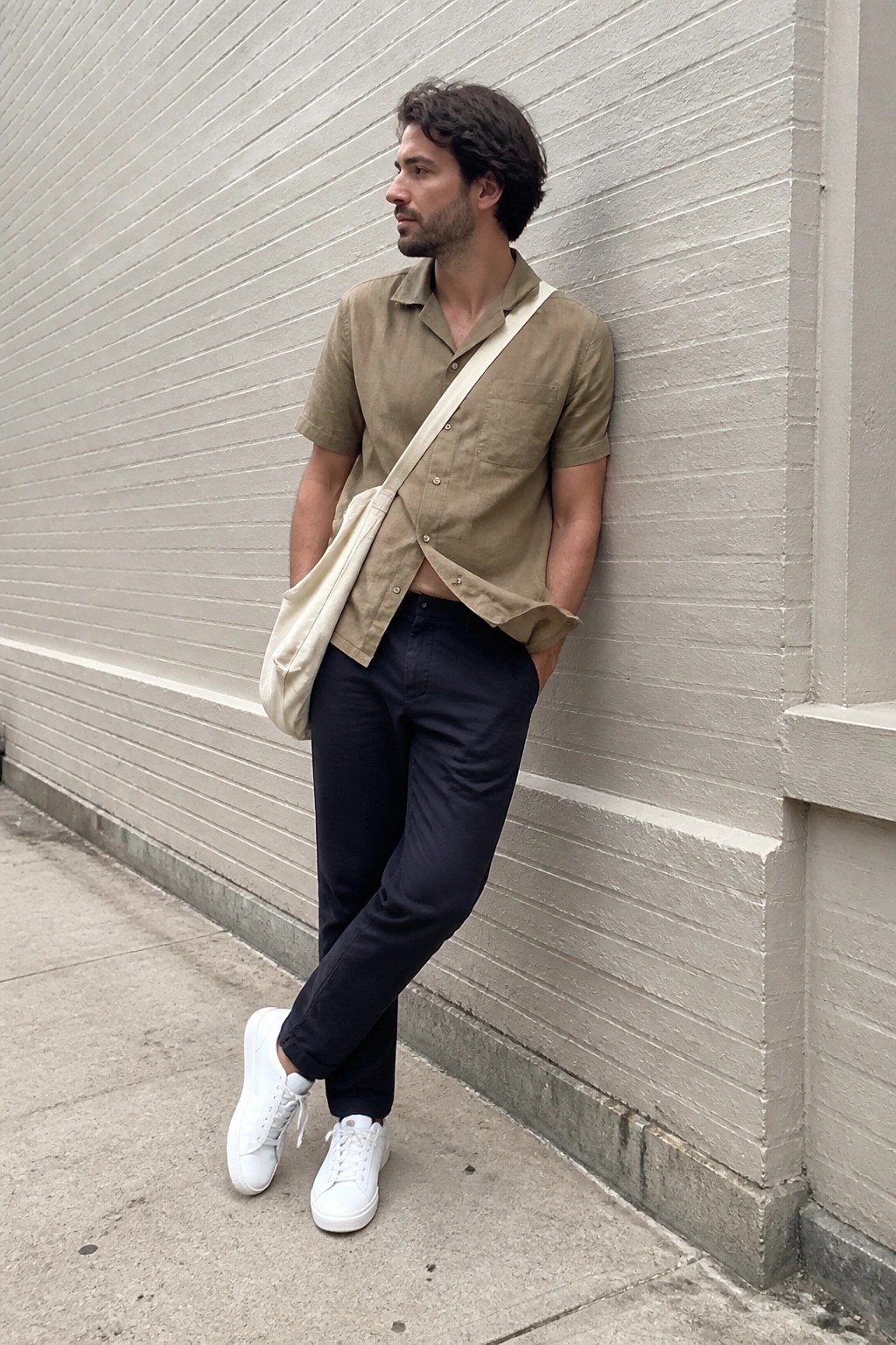 Man in beige shirt and navy pants leaning on wall with crossbody bag