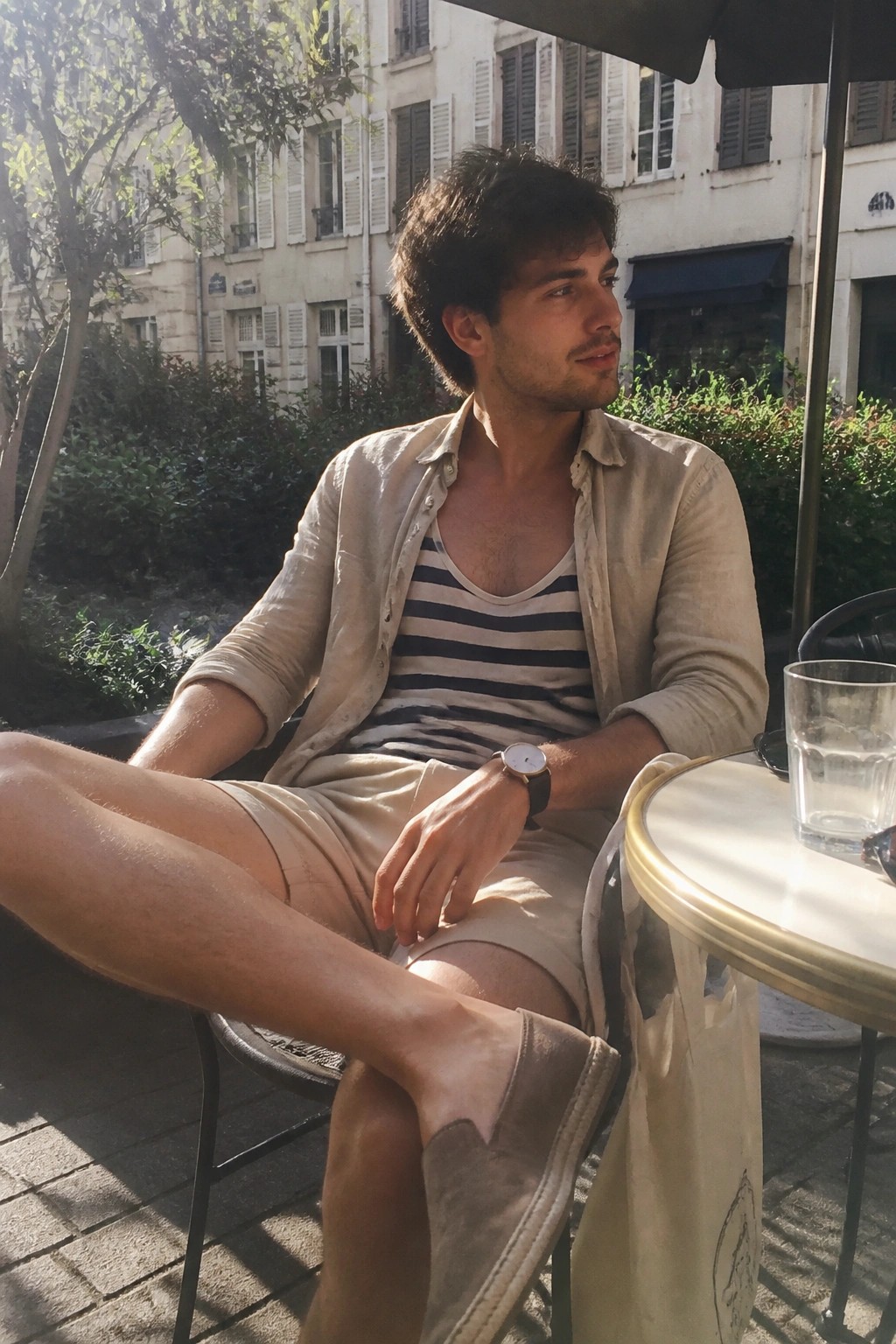 Man in striped tank top and beige shirt at sunny outdoor cafe table.
