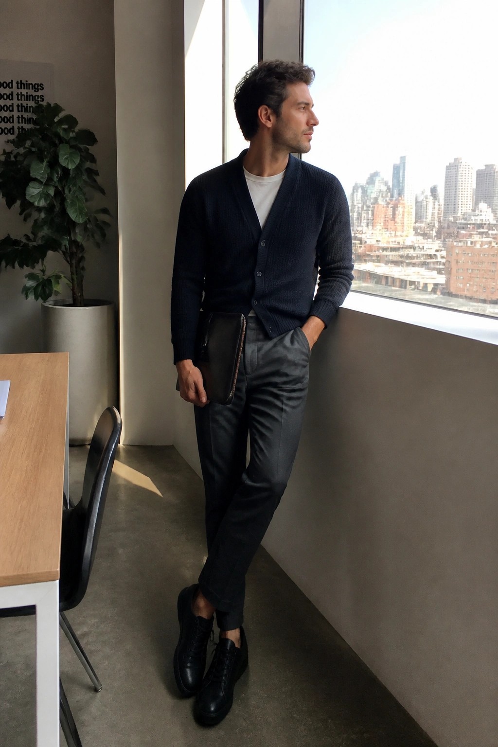 Man in navy cardigan and gray pants holding leather clutch by city window.