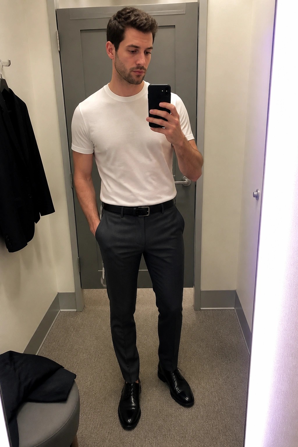 Man in white t-shirt and gray trousers taking mirror selfie in fitting room.